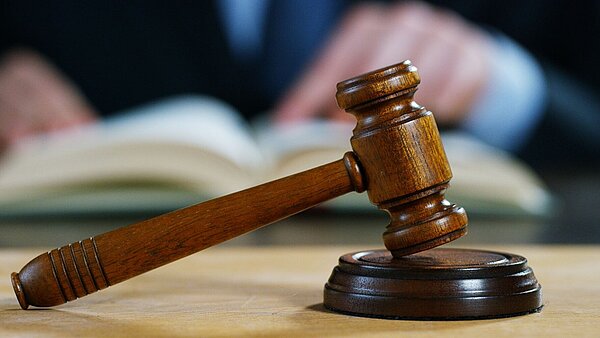 ein Richterhammer in Großaufnahme, im Hintergrund unscharf ein aufgeschlagenes Buch