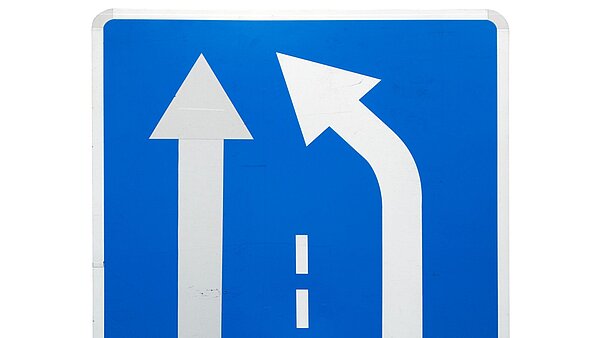 Ein blaues Straßenschild mit weißen Pfeilen, bei dem der eine Pfeil auf den anderen weist