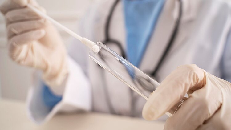 Ein Watteträger wird in ein Röhrchen geschoben von einer unscharf im Hintergrund gezeigten Ärztein